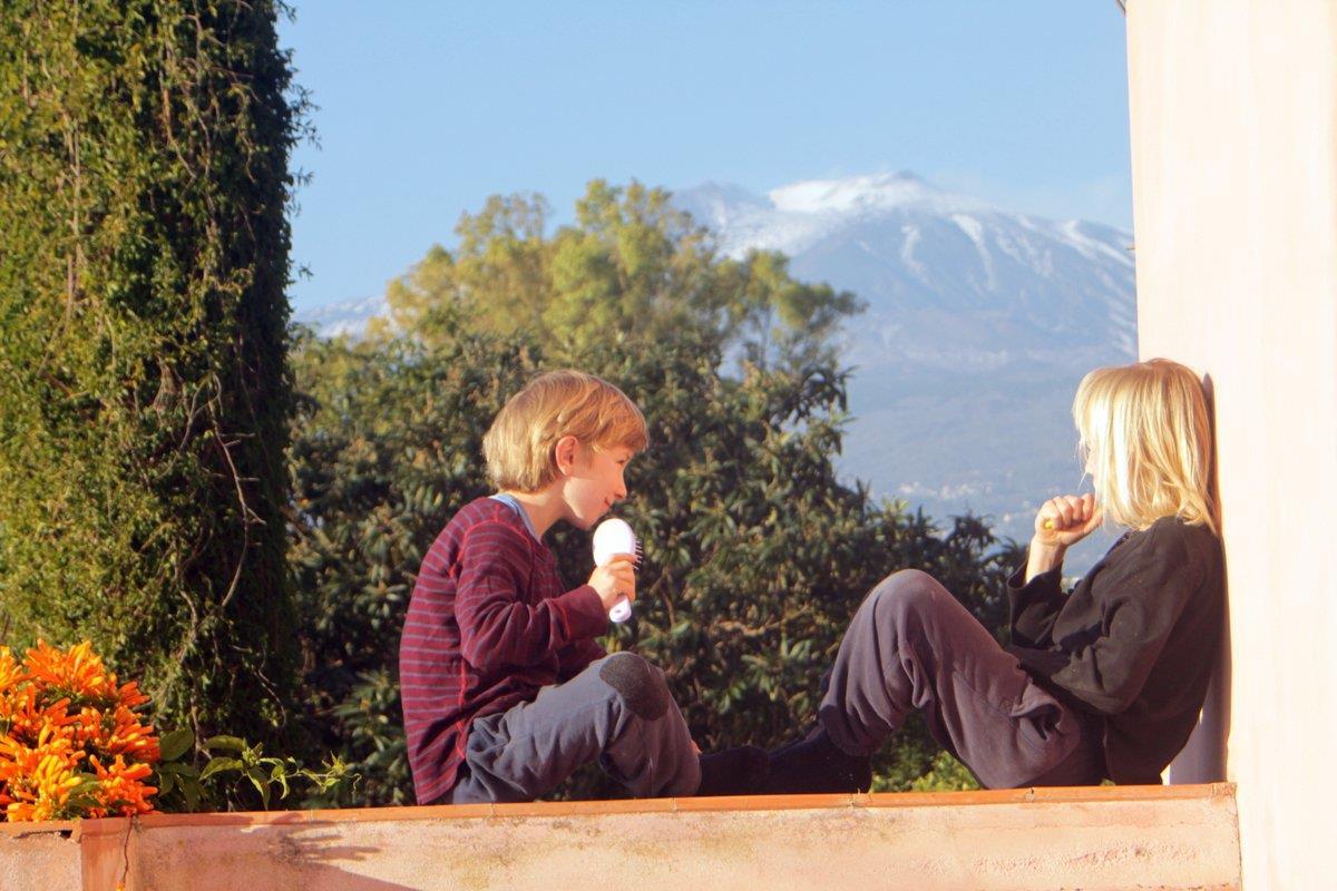 Två barn sitter på ett brett balkongräcke, det är grönt och man ser Etna i bakkrunden